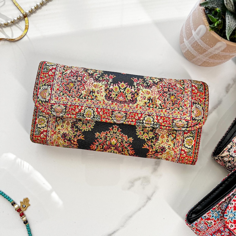 May include: A rectangular clutch with a black background and a detailed red, yellow, and green floral pattern. The clutch has a fold-over flap and appears to be made of a woven fabric. The design resembles a traditional rug.