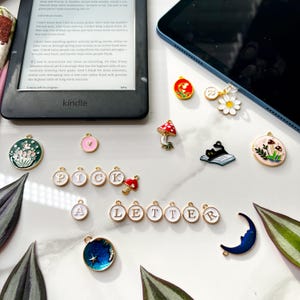 May include: A collection of enamel charms featuring various designs, including a mushroom, a cat, a flower, a moon, and letters spelling out "PICK A LETTER". The charms are gold-toned and have white enamel accents.
