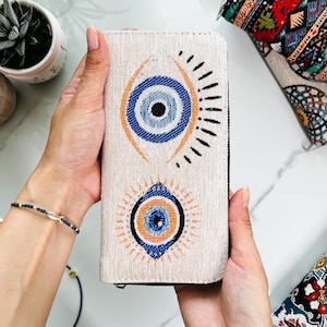 Puede incluir: Una cartera rectangular de color beige con textura de tela tejida y dos motivos prominentes de ojo turco. Cada dise&ntilde;o de ojo consta de c&iacute;rculos conc&eacute;ntricos en azul, naranja y negro, con l&iacute;neas radiantes que se extienden desde los ojos.