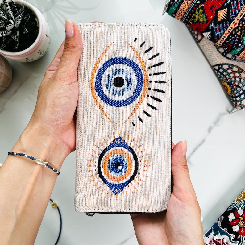 Puede incluir: Una cartera rectangular de color beige con textura de tela tejida y dos motivos prominentes de ojo turco. Cada dise&ntilde;o de ojo consta de c&iacute;rculos conc&eacute;ntricos en azul, naranja y negro, con l&iacute;neas radiantes que se extienden desde los ojos.
