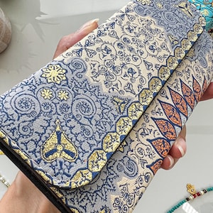 May include: A patterned wallet with a fold-over flap. The wallet features a mix of blue, gold, and cream colors with intricate floral and geometric designs. The edges are trimmed with gold accents, and the wallet appears to be made of a textured fabric.