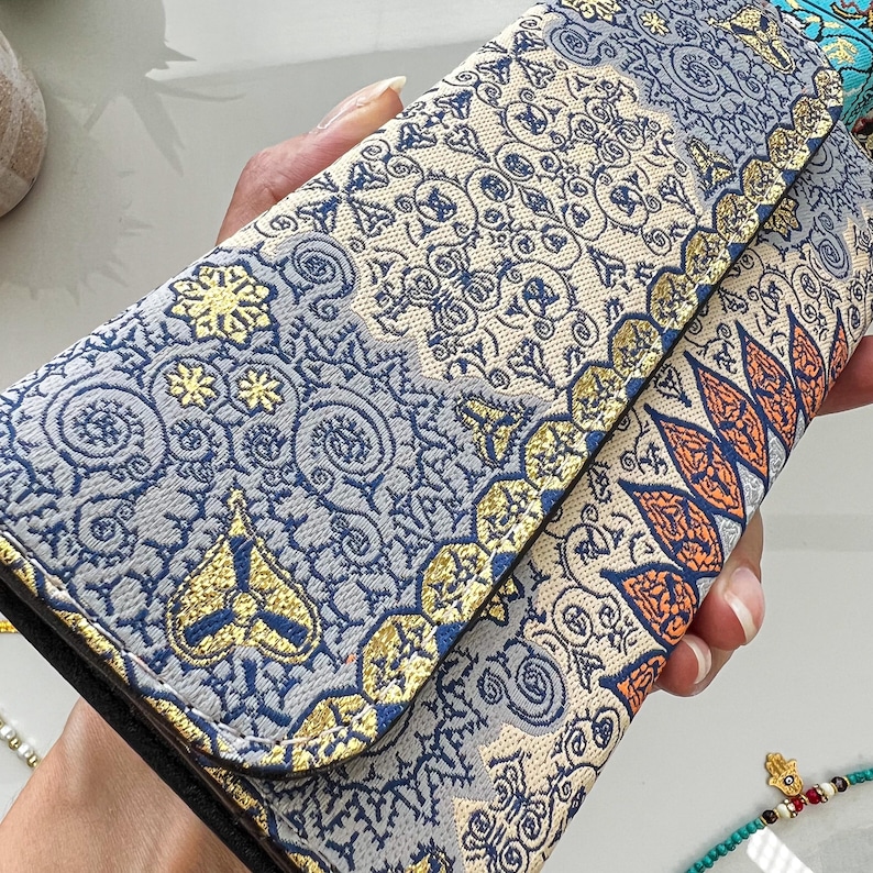 May include: A patterned wallet with a fold-over flap. The wallet features a mix of blue, gold, and cream colors with intricate floral and geometric designs. The edges are trimmed with gold accents, and the wallet appears to be made of a textured fabric.