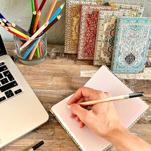 May include: A hand writes in a lined notebook with a pencil. The notebook has a gold patterned cover. A laptop computer, a glass of colored pencils, and five decorative notebooks with patterned covers are on a wooden table.