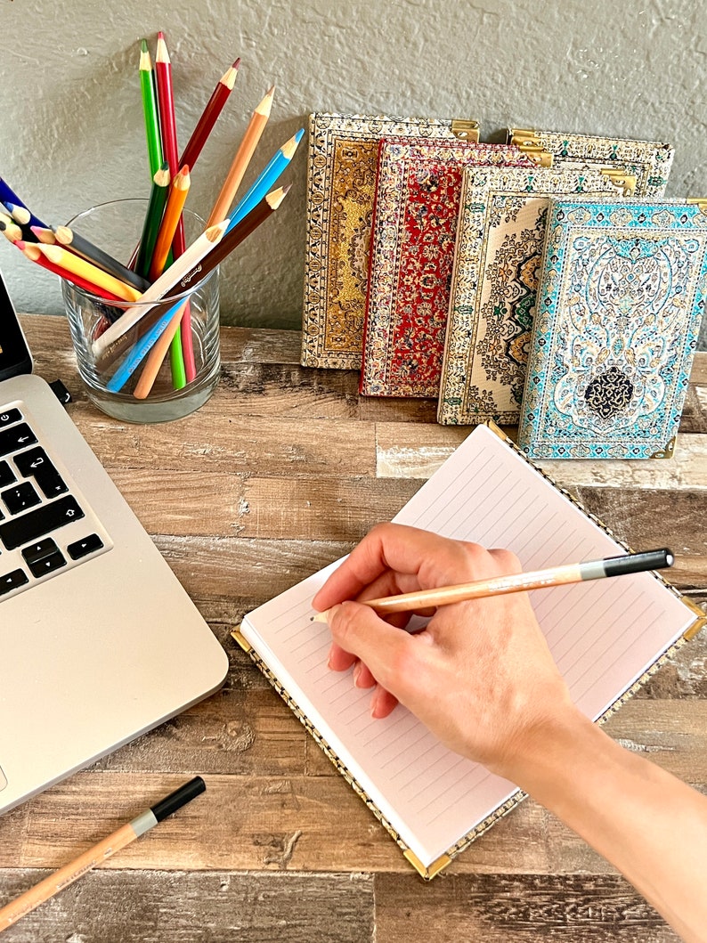 May include: A hand writes in a lined notebook with a pencil. The notebook has a gold patterned cover. A laptop computer, a glass of colored pencils, and five decorative notebooks with patterned covers are on a wooden table.