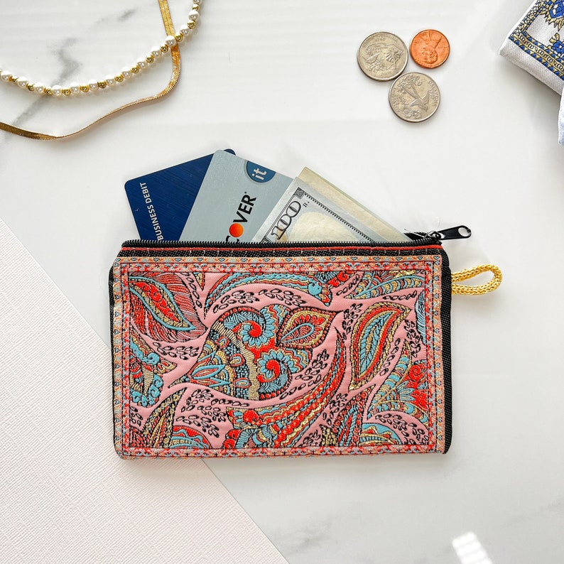 May include: A pink paisley patterned zippered pouch with a black zipper and a yellow pull tab. The pouch is open and contains a credit card, a Discover card, and a dollar bill.