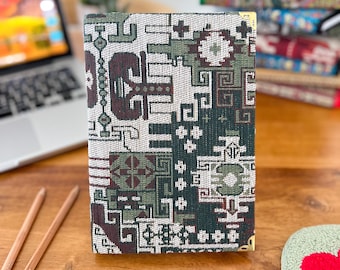Puede incluir: Un cuaderno rectangular con una cubierta de tela tejida con un patrón geométrico en tonos verde oscuro, crema y granate. El cuaderno tiene protectores de esquina de metal dorado en los bordes superior e inferior derecho de la portada.