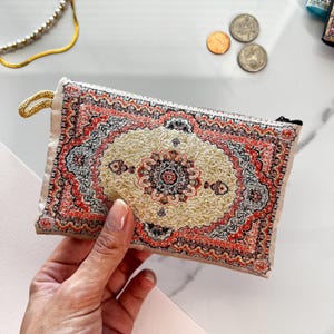 May include: A small zippered pouch with a colorful rug design. The pouch is made of a soft, beige fabric and has a zipper closure. The rug design features intricate patterns in shades of red, gold, and black.