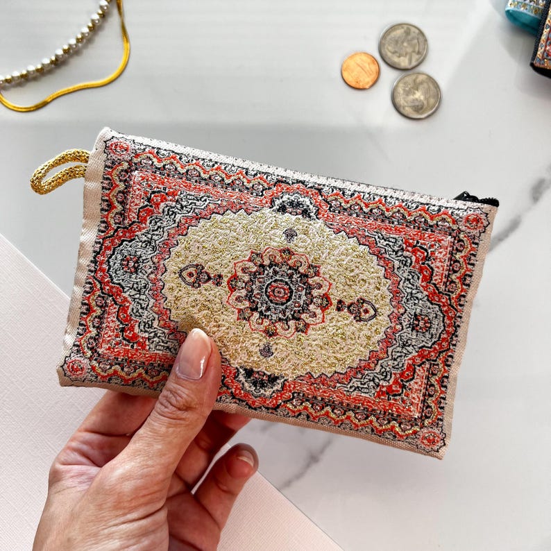 May include: A small zippered pouch with a colorful rug design. The pouch is made of a soft, beige fabric and has a zipper closure. The rug design features intricate patterns in shades of red, gold, and black.