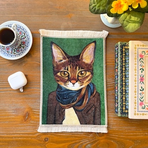 May include: A rectangular green rug with a cat portrait wearing a blue scarf and brown jacket. The cat has brown and gold fur against a green backdrop. The rug has a fringed edge, and is likely a decorative item.