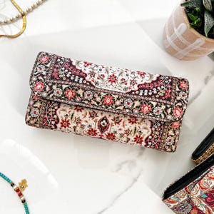May include: A rectangular clutch with a detailed floral pattern in red, black, and white. The clutch has a fold-over flap and appears to be made of a textured fabric. The design is reminiscent of a traditional rug.