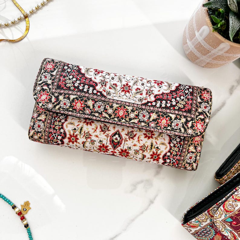 May include: A rectangular clutch with a detailed floral pattern in red, black, and white. The clutch has a fold-over flap and appears to be made of a textured fabric. The design is reminiscent of a traditional rug.