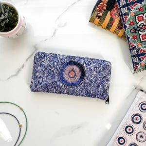 Puede incluir: Una cartera rectangular con un patr&oacute;n abstracto texturizado en azul y blanco con un motivo circular prominente en el centro. La cartera tiene un cierre de cremallera negro en el borde superior y est&aacute; hecha de tela tejida.