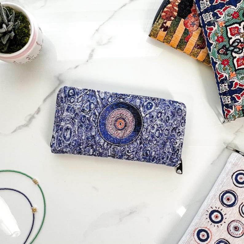 Puede incluir: Una cartera rectangular con un patr&oacute;n abstracto texturizado en azul y blanco con un motivo circular prominente en el centro. La cartera tiene un cierre de cremallera negro en el borde superior y est&aacute; hecha de tela tejida.