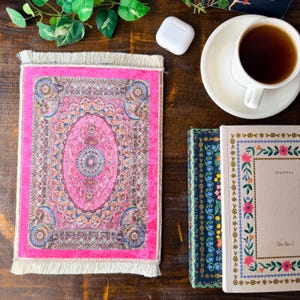 May include: A rectangular coaster featuring a vibrant pink and multi-colored intricate oriental rug pattern with a central medallion, ornate floral borders, and cream-colored fringe trim on the top and bottom edges of the fabric surface.