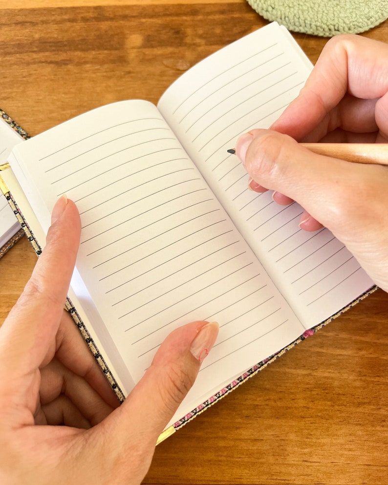 May include: A small, lined notebook with a patterned cover in shades of pink, black, and gold. The notebook is open and a hand is writing with a pencil.