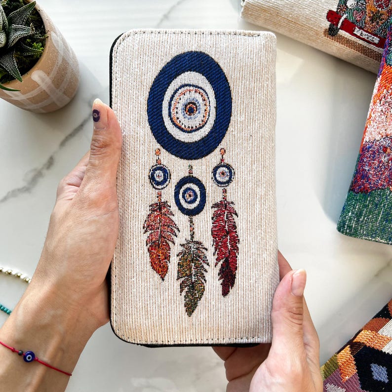 Puede incluir: Una cartera rectangular con exterior de tela texturizada en color blanco roto que presenta un dise&ntilde;o tejido de un gran s&iacute;mbolo de ojo turco azul oscuro en la parte superior, con tres amuletos de ojo m&aacute;s peque&ntilde;os colgando debajo, cada uno con una pluma roja y verde.