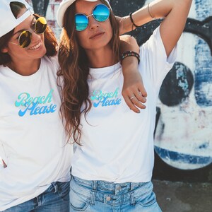 May include: Two women wearing white t-shirts with the text "Beach Please" in a blue and green gradient. They are both wearing sunglasses and smiling.