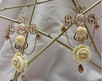 May include: A pair of gold tone earrings with a cross design and white rose accents. Each earring features a cameo with a woman's profile and a dangling crystal.