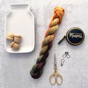 May include: A white ceramic mason jar tray containing three small wooden acorn shapes, a skein of variegated yarn in orange, yellow, and green, a black circular measuring tape with the text Love Beyond Measure, gold scissors, and two black stitch markers.