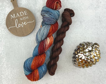 May include: Two skeins of yarn, one variegated in blue and rust orange and one solid dark brown, alongside a round wooden tag with the text MADE WITH love and a metallic silver pinecone-shaped ornament with a textured, reflective surface.