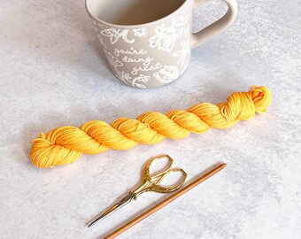 Peut inclure: Une pelote de laine jaune, une paire de ciseaux dorés et une aiguille à tricoter en bois sur une surface blanche. Une tasse avec le texte "you're doing great" est en arrière-plan.