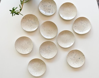 May include: Ten small, white ceramic dishes with a speckled pattern. The dishes are arranged in a circle on a white surface.