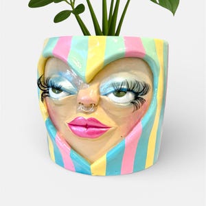 May include: A colorful ceramic planter featuring a stylized face with long eyelashes, a nose ring, and pink lips. The planter is decorated with vertical stripes in pastel colors and holds a green plant.