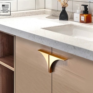 May include: Gold cabinet hardware with a triangular shape. The hardware is mounted on a light brown cabinet door.