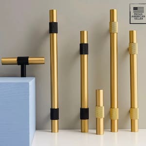 Könnte beinhalten: Eine Sammlung goldfarbener Schrankgriffe und -knöpfe mit schwarzen und strukturierten Akzenten. Die Hardware wird vor einem neutralen Hintergrund präsentiert und zeigt verschiedene Größen und Designs. Das Bild enthält einen "Proud American Seller"-Aufkleber.