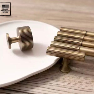May include: A set of five brass cabinet pulls with a textured, ribbed design. The pulls are arranged in a row on a white plate. The pulls have a T-shaped design with a round knob on the end. The pulls are a brushed brass finish.