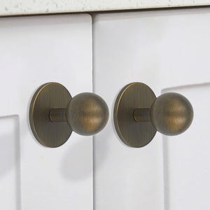 May include: Two antique brass cabinet knobs with round, slightly raised, ball-shaped handles. The knobs are mounted on white cabinet doors.