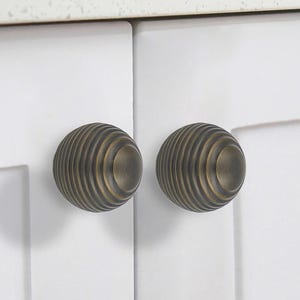 May include: Two antique brass cabinet knobs with a ribbed design. The knobs are mounted on white cabinet doors. The text "DESIGN MAX" is printed on the bottom right corner of the image.
