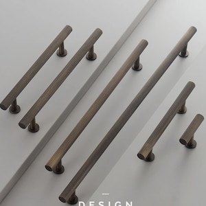 May include: A set of seven brushed brass cabinet pulls in various sizes. The pulls have a ridged texture and a round shape. The pulls are arranged in a staggered pattern on a white surface. DESIGN MAX is printed in black text below the pulls.