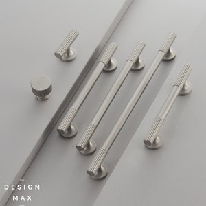 以下が含まれることがあります： 6つのブラシ仕上げニッケル製のキャビネット金具のプルとノブのセット。プルは長く、細く、リブ模様になっています。ノブは丸く、同様のリブ模様になっています。金具はグレーの背景に配置されています。