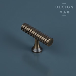 May include: A brushed antique brass cabinet pull with a T-shaped design. The pull has a cylindrical handle with vertical grooves and a round base.