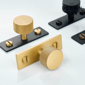 Può includere: Due pomelli per armadi dorati con una superficie testurizzata. I pomelli sono montati su piastre rettangolari nere con viti. Le piastre sono su una superficie bianca. Il testo "DESIGN MAX" è visibile su una delle piastre.