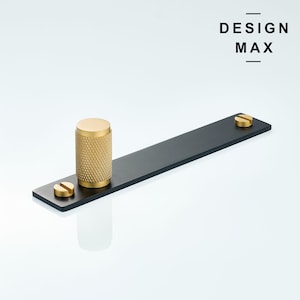 Op de afbeelding: Zwarte metalen ladegreep met een gouden knop en twee gouden schroeven.  DESIGN MAX is gedrukt in zwarte tekst op een witte achtergrond.