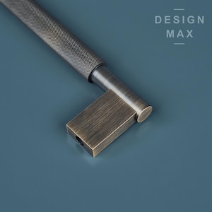 May include: A brushed nickel cabinet pull with a square, antique brass end cap. The pull has a textured, ridged surface.  DESIGN MAX is printed in white on a blue background.
