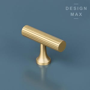 Puede incluir: Un pomo de armario en forma de T de color dorado con un diseño acanalado.  DESIGN MAX está impreso en la esquina superior derecha de la imagen.