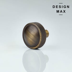 May include: A bronze cabinet knob with a textured rim and a smooth, domed centre. The knob has a short, cylindrical stem. The text "DESIGN MAX" is displayed in black, above the knob.