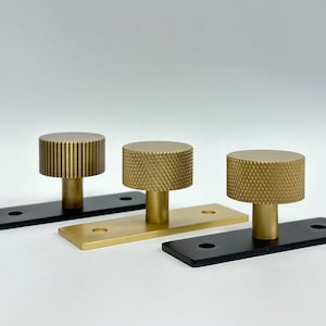 May include: Three gold-toned cabinet knobs with different patterns. The knobs are mounted on black and gold rectangular bases.