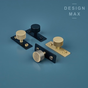 May include: Three cabinet knobs with a black and brass finish. The knobs are round with a textured design and are mounted on black rectangular plates. The plates have two screws each.