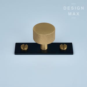 Può includere: Una manopola per armadio dorata con una superficie testurizzata e una piastra di base nera. La manopola è fissata alla piastra di base con due viti. Il testo "DESIGN MAX" è stampato in bianco su sfondo nero.