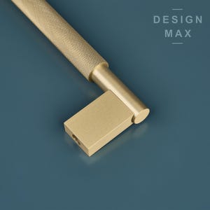 Pode incluir: Uma puxador de armário dourado com uma pega cilíndrica texturizada e uma base quadrada. O puxador está sobre um fundo azul.