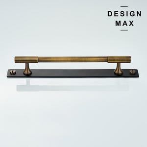May include: A brushed brass pull handle with a black rectangular base and two screws. The handle has a textured design and is mounted on a white background. The text "DESIGN MAX" is visible in the top right corner of the image.