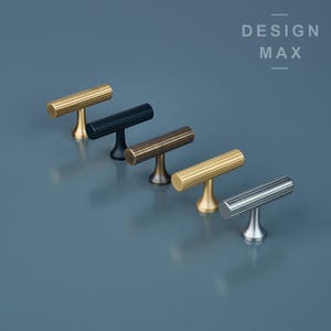 May include: Five different finishes of a t-bar cabinet pull. The pulls are arranged in a row from left to right, starting with a gold finish, then black, bronze, brass, and finally silver.