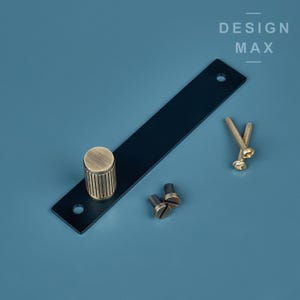 Puede incluir: Un tirador de cajón de metal negro con un pomo de latón y dos tornillos. El tirador está sobre un fondo azul con el texto "DESIGN MAX" encima.