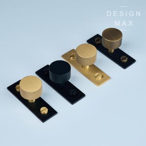 Könnte beinhalten: Vier Schrankknöpfe, zwei goldfarben und zwei schwarz, mit einem strukturierten Design. Jeder Knopf ist auf einer schwarzen rechteckigen Platte mit Schrauben montiert.