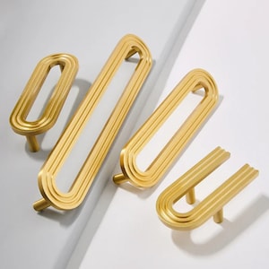 May include: Four gold-toned metal cabinet pulls in varying lengths, each featuring an elongated oval or U-shaped design with a ribbed, stepped surface texture and two cylindrical mounting posts for attachment to furniture or cabinetry doors.