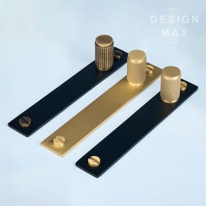 May include: Three brass cabinet knobs with different patterns, each mounted on a black or gold metal bar. The bars are labeled "DESIGN MAX".